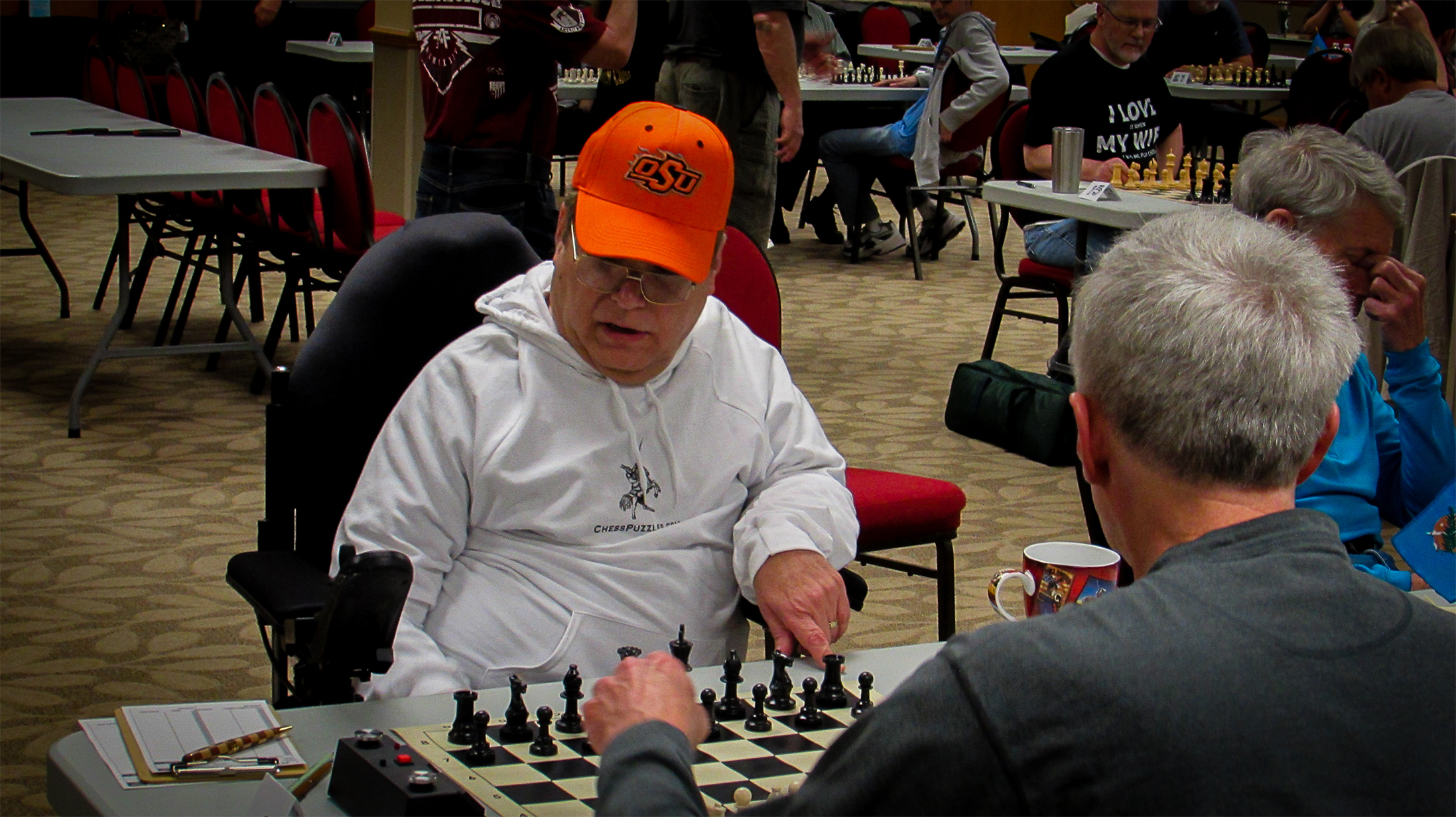Wayne Hatcher (wearing the orange hat) split his match on Board 2.  He is ranked in the 95th Percentile for all USA players and in the 90th Percentile for all USA Seniors.  He is ranked 54th in Blitz for all USA Seniors.  He is ranked Number 19 for all Oklahoma players.  In 2009 he won the 26-player Arizona Senior Open with a 4.5/5.0 score.  Photo by Mike Tubbs.